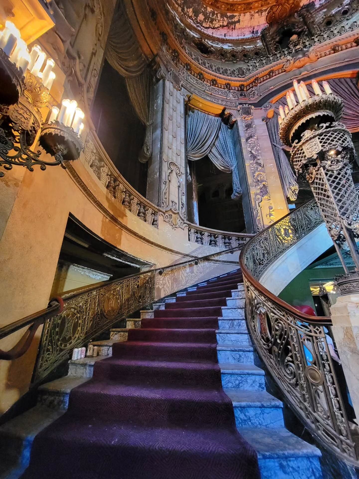 A carpeted marble staircase with ornate iron railings curves up through the Uptown Theatre's grand lobby, lit by candelabra-style lamps.