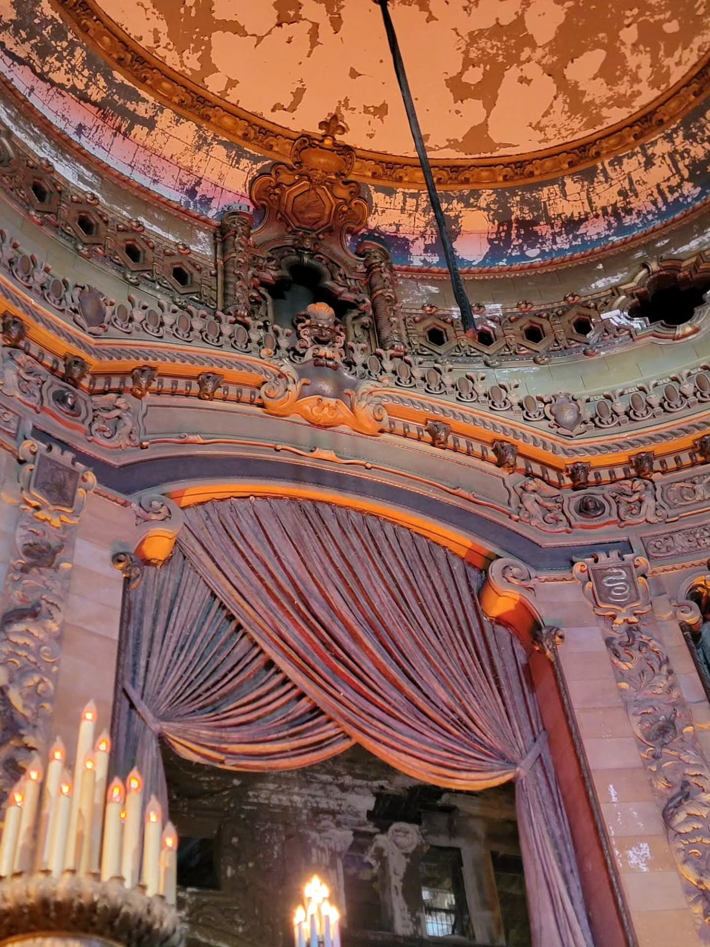 Peeling paint on an ornate domed ceiling above a draped arch and candelabra lamps inside the Uptown Theatre.